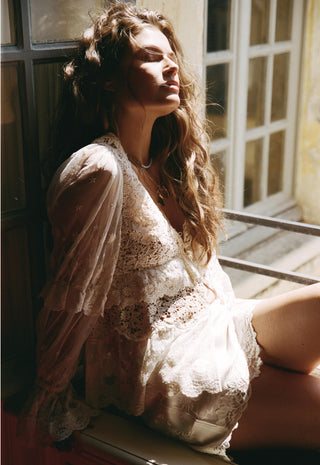 a-woman-with-long-wavy-brown-hair-sits-by-a-sunlit-window-wearing-a-sheer-cream-lace-robe-and-matching-lace-lingerie-the-warm-sunlight-creates-a-soft-dreamy-look-with-strong-shadows-across-her-face-and-body