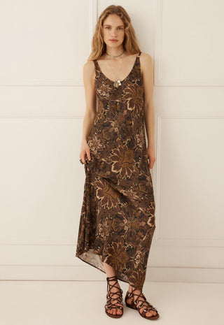a-woman-modeling-a-sleeveless-brown-maxi-dress-with-a-metallic-bronze-floral-pattern-paired-with-brown-strappy-sandals-standing-against-a-white-paneled-wall