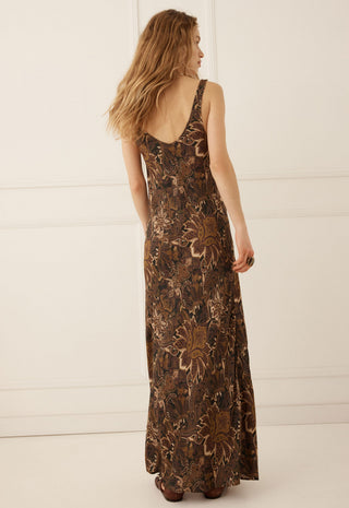 a-woman-wearing-a-sleeveless-floor-length-brown-maxi-dress-with-a-floral-pattern-shown-from-the-back-against-a-white-paneled-wall