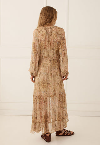 A woman viewed from behind wearing a long-sleeved, midi-length beige maxi dress with an intricate paisley pattern in warm brown and tan tones. She is wearing brown strappy flat sandals against a clean, white paneled wall background.
