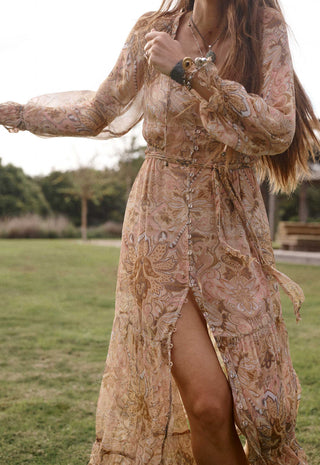 A woman outdoors in a grassy park wearing a long, flowing boho-style maxi dress with a high slit. The dress has an intricate paisley and floral pattern in muted beige, tan, and brown tones with subtle gold and blue accents.
