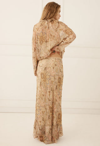 a-woman-stands-with-her-back-to-the-camera-wearing-a-matching-two-piece-set-consisting-of-a-long-sleeve-cropped-top-and-a-long-skirt-both-in-a-beige-and-brown-paisley-patterned-fabric