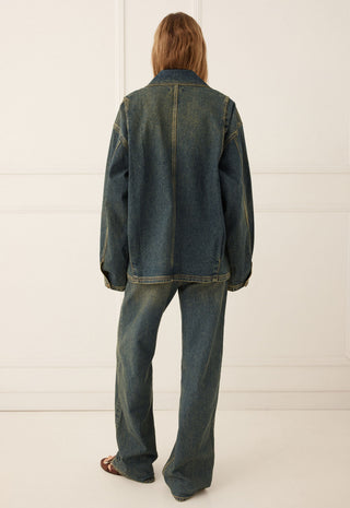 a-model-viewed-from-the-back-wearing-an-oversized-dark-blue-denim-jacket-and-matching-wide-leg-denim-jeans-standing-against-a-white-paneled-wall