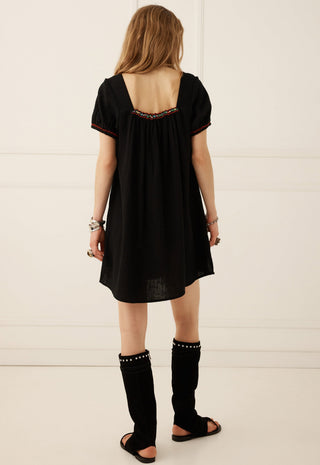 a-woman-viewed-from-behind-wearing-a-loose-black-short-sleeve-dress-with-a-decorative-embroidered-neckline-black-studded-leg-warmers-and-black-sandals-standing-against-a-white-paneled-wall