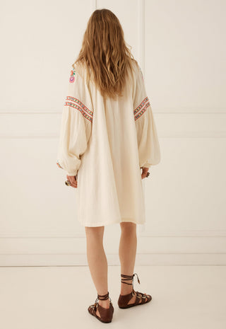a-woman-viewed-from-behind-wearing-a-cream-colored-bohemian-long-sleeve-tunic-dress-with-red-and-orange-geometric-embroidery-on-the-sleeves-paired-with-brown-strappy-sandals-set-against-a-white-paneled-wall