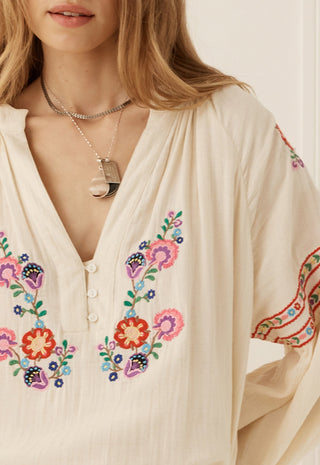 a-close-up-of-a-woman-wearing-a-cream-colored-bohemian-blouse-with-colorful-floral-embroidery-on-the-front-and-sleeves-accessorized-with-layered-silver-necklaces