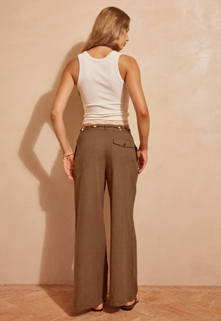 a-woman-shown-from-the-back-wearing-wide-leg-brown-trousers-and-a-white-ribbed-tank-top-standing-against-a-textured-beige-wall