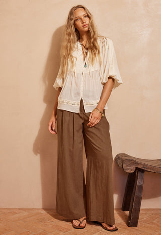a-woman-stands-in-a-warm-toned-room-wearing-a-loose-white-blouse-with-lace-trim-and-wide-leg-brown-pants-paired-with-sandals-a-rustic-stool-is-visible-against-a-textured-beige-wall