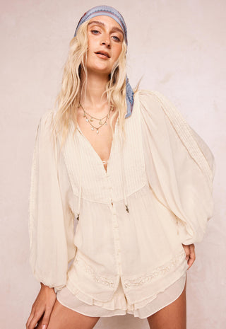 a-woman-modeling-a-cream-colored-long-sleeved-bohemian-style-romper-with-pleated-details-and-lace-trim-paired-with-a-light-blue-patterned-headband-against-a-neutral-beige-background