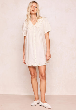 a-woman-modeling-a-short-sleeve-cream-colored-mini-dress-with-a-ruffled-peter-pan-collar-and-eyelet-lace-trim-paired-with-white-lace-up-flats-against-a-textured-beige-wall