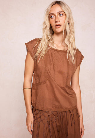 a-woman-with-long-blonde-hair-wearing-a-brown-sleeveless-top-and-matching-brown-skirt-standing-against-a-textured-light-beige-background
