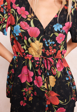 a-close-up-of-a-black-short-sleeve-v-neck-dress-with-a-vibrant-floral-print-featuring-pink-red-blue-and-yellow-flowers-with-green-leaves-the-dress-has-a-drawstring-waist-and-button-details-down-the-front