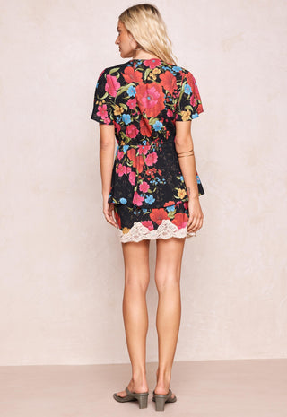 a-woman-viewed-from-the-back-wearing-a-short-sleeve-black-dress-with-a-vibrant-multicolor-floral-print-and-a-white-lace-hem-paired-with-gray-block-heel-sandals-against-a-textured-beige-wall