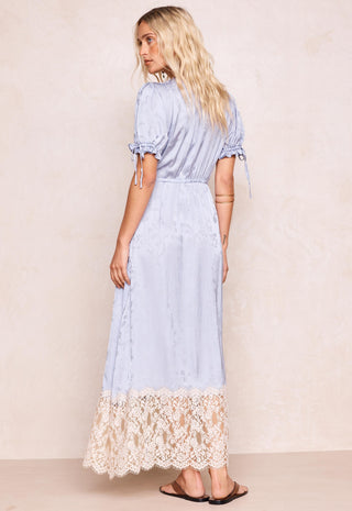 a-woman-modeling-a-light-blue-maxi-dress-with-short-puff-sleeves-side-tie-details-and-a-cream-lace-hem-shown-from-the-back-against-a-textured-beige-wall