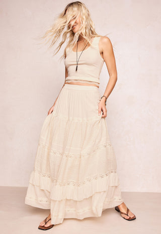 a-woman-modeling-a-cream-colored-two-piece-outfit-consisting-of-a-sleeveless-crop-top-and-a-long-tiered-maxi-skirt-with-delicate-lace-trim-paired-with-brown-flip-flop-sandals-against-a-textured-beige-studio-backdrop