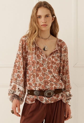 a-woman-modeling-a-long-sleeve-bohemian-style-blouse-with-a-rust-brown-and-cream-floral-pattern-paired-with-a-dark-brown-belt-and-brown-pants-set-against-a-white-paneled-wall
