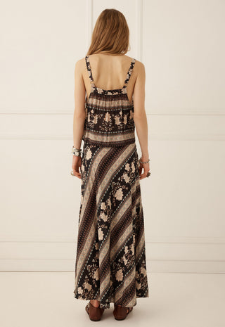 a-woman-viewed-from-behind-wearing-a-sleeveless-maxi-dress-with-a-brown-beige-and-black-striped-and-floral-pattern-paired-with-brown-flat-shoes-against-a-white-paneled-wall