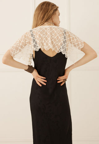 a-woman-viewed-from-the-back-wearing-a-black-slip-dress-with-a-sheer-white-lace-flutter-cape-overlay-set-against-a-light-cream-paneled-wall