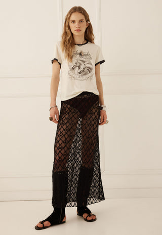 a-model-stands-against-a-white-paneled-wall-wearing-a-cream-graphic-t-shirt-with-black-trim-sheer-black-patterned-maxi-pants-and-black-sandals