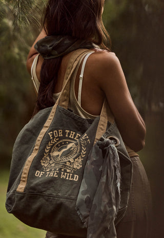 a-woman-outdoors-carrying-a-large-canvas-tote-bag-over-her-shoulder-the-bag-is-dark-olive-green-with-tan-straps-and-a-tan-graphic-print-that-reads-for-the-love-of-the-wild-around-a-illustration