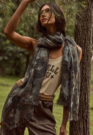 a-woman-outdoors-in-a-forest-setting-wearing-a-lightweight-camouflage-scarf-with-fringed-edges-over-a-beige-tank-top-and-brown-pants