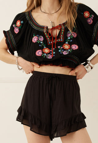 A woman wearing a black embroidered crop top with colorful floral patterns and matching black ruffled shorts, standing against a light beige wall.